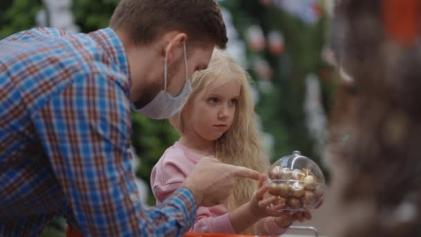 Батько в медичній масці на обличчі зі своєю дочкою вибирає ювелірні вироби та іграшки напередодні Різдва, щоб прикрасити будинок і ялинку . — стокове відео