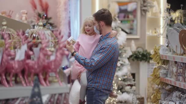Ein Vater mit medizinischer Maske im Gesicht und seine Tochter suchen sich an Heiligabend Schmuck und Spielzeug aus, um Haus und Weihnachtsbaum in Zeitlupe zu schmücken. — Stockvideo
