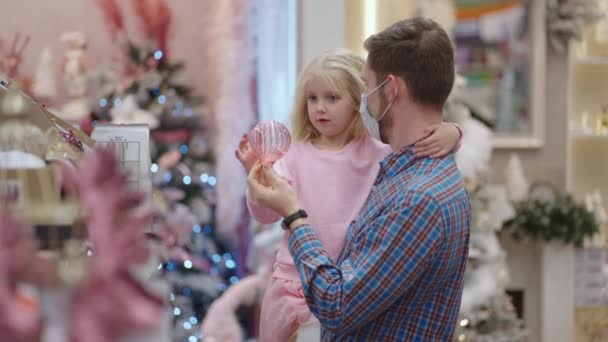 Ein Vater mit medizinischer Maske im Gesicht und seine Tochter suchen sich an Heiligabend Schmuck und Spielzeug aus, um Haus und Weihnachtsbaum zu schmücken — Stockvideo