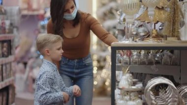 Oğluyla birlikte tıbbi maske takan bir anne Noel için ev dekorasyonunu seçer. Noel ağacı oyuncaklarını izle. Coronavirüs salgını. Dekorasyon ve dekorasyonları seç