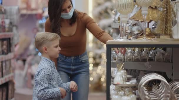 モールで息子と一緒に医療マスクの母親はクリスマスのための家の装飾を選択します。クリスマスツリーのおもちゃを見てください。コロナウイルスの流行。装飾や装飾を選択します — ストック動画