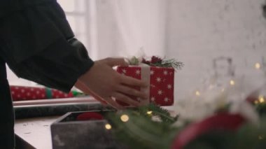 Tahta bir masada çelenklerle hediye süsleyen genç bir kadın döndü ve masaya hazır yapılmış bir Noel kutusunu kurdeleyle ezdi..