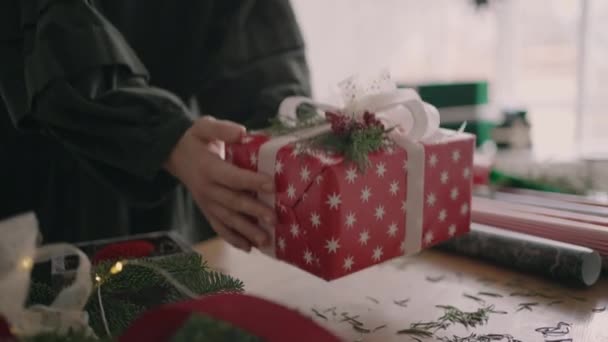 Молодая женщина украшает подарок на деревянном столе с гирляндами переворачивается и кладет на стол готовую рождественскую коробку, раздавленную лентой в замедленной съемке — стоковое видео