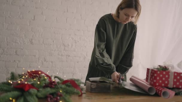 Portrait Une jeune femme décorant un cadeau sur une table en bois avec des guirlandes se retourne et pose sur la table une boîte de Noël prête à l'emploi broyée avec un ruban — Video