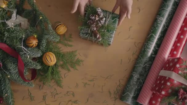 Plan de vue de dessus. Entièrement visible la table avec les décorations. mains mettre et finaliser cadeau de Noël enveloppé dans du papier artisanat sur une table en bois. Ruban de bandage et noeud noué. — Video