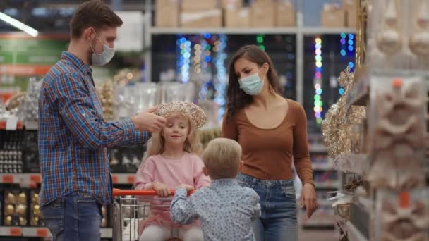 Ένα παντρεμένο ζευγάρι με δύο παιδιά σε ένα εμπορικό κέντρο με προστατευτικές μάσκες στην επιδημία του κορωναϊού ετοιμάζονται για τα Χριστούγεννα και επιλέγουν διακοσμήσεις. — Αρχείο Βίντεο