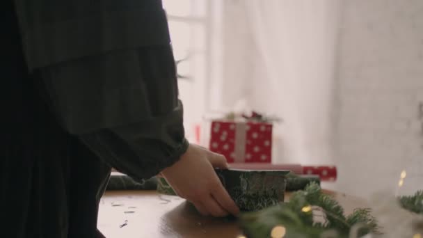 Vista lateral de montar y preparar un regalo de Navidad. Un primer plano de una mujer envuelve sus manos alrededor de una caja en papel de regalo con un adorno navideño. envoltura de regalo decorador profesional — Vídeo de stock