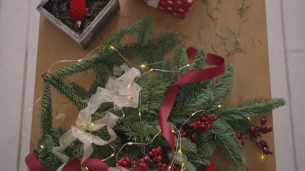 Plan de vue de dessus. Entièrement visible la table avec les décorations. Les mains féminines mettent et finalisent le cadeau de Noël enveloppé dans du papier artisanal sur une table en bois. Ruban de bandage et noeud noué. — Video
