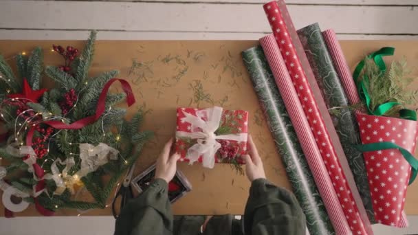 Plan du haut vers le bas : Une femme met un cadeau sur la table pour l'emballage et resserre un ruban cadeau dessus. Décorateur et table d'emballage cadeau — Video