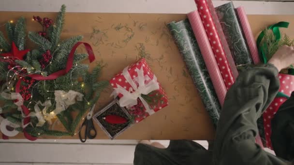 Un plan descendant. Entièrement visible la table avec les décorations. Les mains féminines mettent et finalisent le cadeau de Noël enveloppé dans du papier artisanal sur une table en bois. Bande de bandage — Video