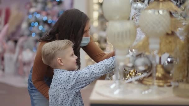 Eine Mutter in medizinischer Maske mit ihrem Sohn in einem Einkaufszentrum wählt Weihnachtsdekoration — Stockvideo