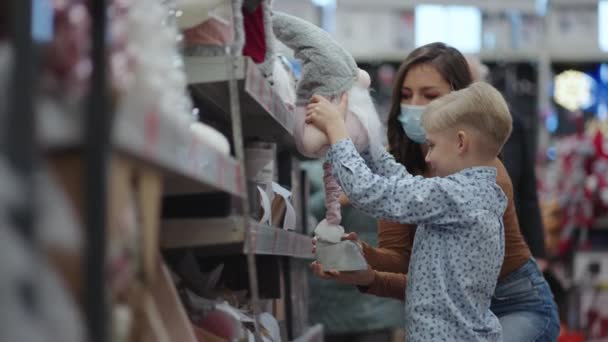 Мама в защитной маске с мальчиком забирает игрушку в торговом центре в канун Рождества во время пандемии — стоковое видео