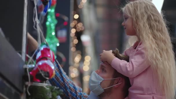 Πατέρας και κόρη κάθονται στους ώμους τους σε ένα εμπορικό κέντρο επιλέγουν μια γιρλάντα για το σπίτι και χριστουγεννιάτικο δέντρο για τα Χριστούγεννα κατά τη διάρκεια της πανδημίας σε αργή κίνηση. — Αρχείο Βίντεο