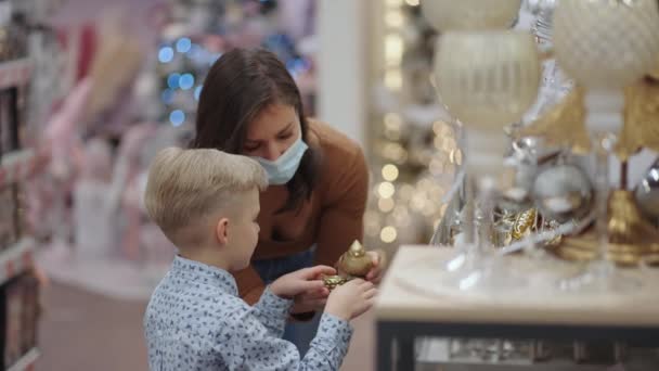 Una madre en una máscara médica con su hijo en el centro comercial elegir decoraciones para el hogar para la Navidad. Nochebuena durante la pandemia — Vídeo de stock