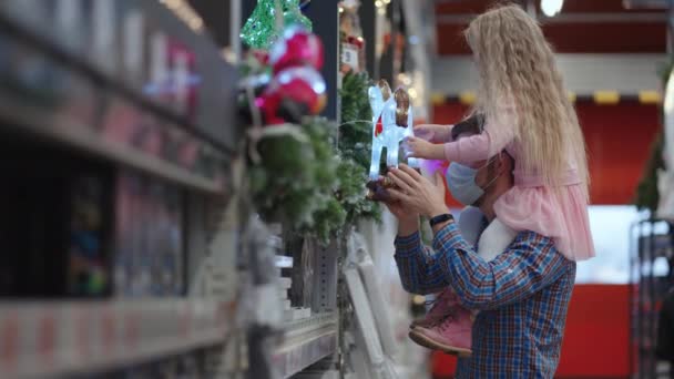 Un padre camina con una máscara protectora alrededor de la tienda con su hija sobre sus hombros en un supermercado y elige un caballo brillante — Vídeos de Stock