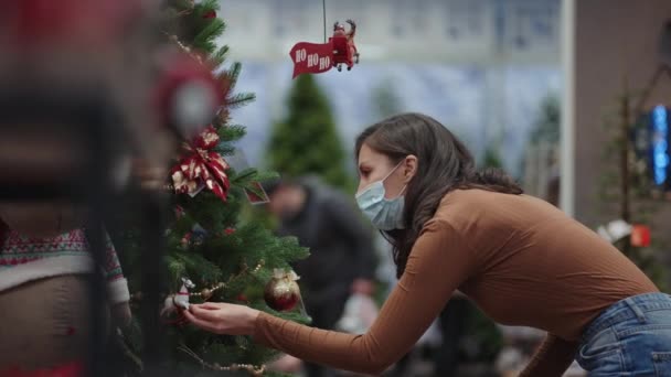 戴着防护面具的女人选择圣诞装饰品。准备圣诞节在大脑皮层的大流行。圣诞树玩具和花环 — 图库视频影像