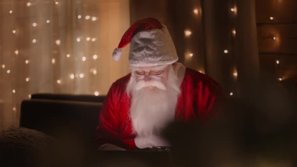 Porträt des Weihnachtsmannes, der nachts mit einem Laptop im Licht der Weihnachtsbeleuchtung arbeitet — Stockvideo