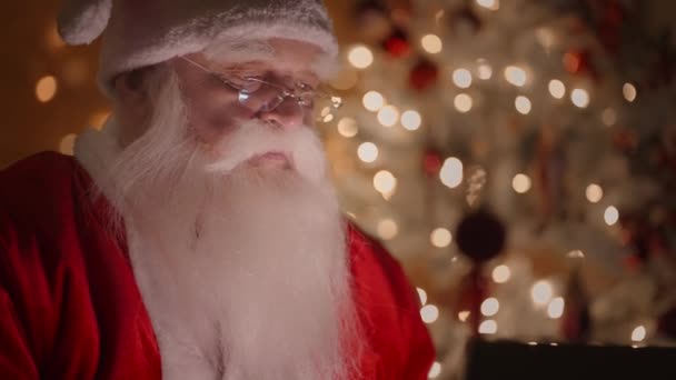 Крупный план реального Санта-Клауса, работающего ночью с ноутбуком в очках в свете рождественских огней на фоне елки. Удаленная работа — стоковое видео
