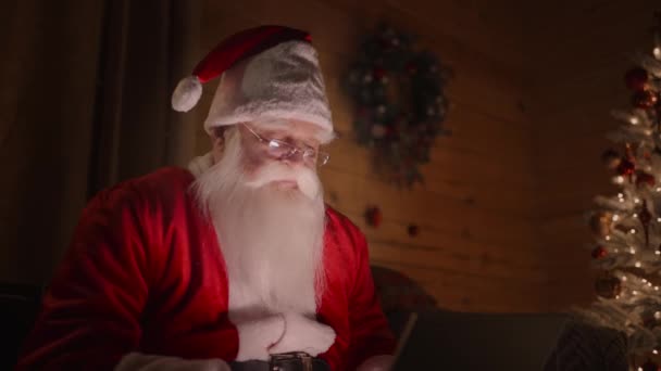 Święty Mikołaj pracuje na komputerze w domu z okularami i brodą magiczne lampki świąteczne na tle ozdobionej choinki — Wideo stockowe