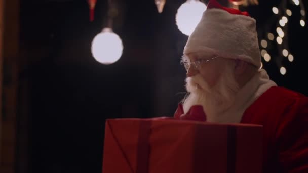 Portret van een echte kerstman met een bril en een baard op de buitenlucht komt naar het raam van het huis met een geschenk op de avond voor Kerstmis en kijkt naar binnen. Kerstman brengt een geschenk — Stockvideo