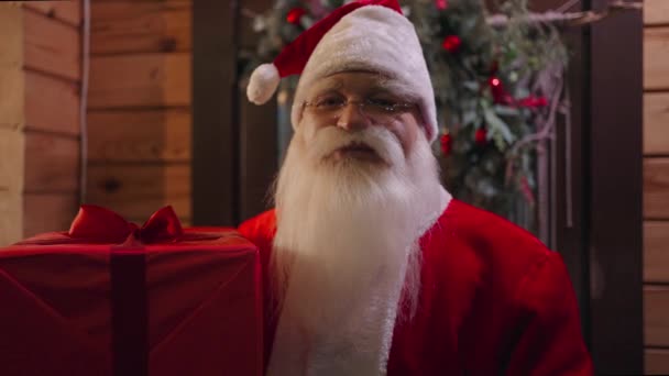 Santa Claus looks into the camera and enters the house to leave gifts under the Christmas tree in winter in December, the camera moves away from the portrait to the general plan — Stock Video