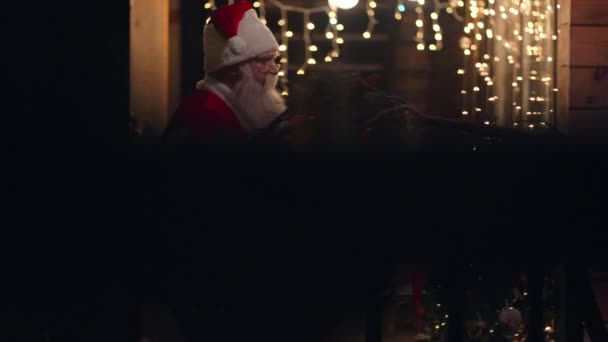 Πλάγια άποψη Άγιος Βασίλης μεταφέρει και παραδίδει ένα δώρο στα χέρια του το χειμώνα στο δρόμο και έρχεται στην πόρτα και μπαίνει στο σπίτι με γιρλάντες και χριστουγεννιάτικα στολίδια. Τον Δεκέμβριο παραμονή Χριστουγέννων — Αρχείο Βίντεο