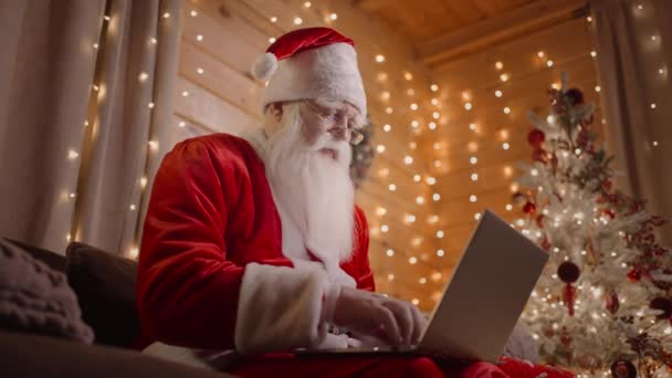 Papai Noel está trabalhando no computador em casa nas luzes de Natal mágicas no fundo de uma árvore de Natal decorada — Vídeo de Stock