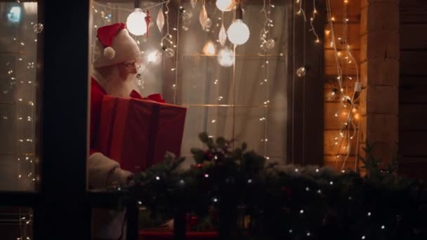 Boční pohled Santa Claus nese a dodává dárek v jeho rukou v zimě na ulici a přichází ke dveřím a vstupuje do domu s věnci a vánoční dekorace. Kouzelný okamžik — Stock video