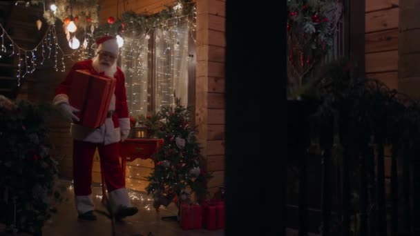 Side view Santa Claus carries and delivers a gift in his hands in winter on the street and comes to the door and enters the house with garlands and Christmas decorations. In December on Christmas Eve — Stock Video