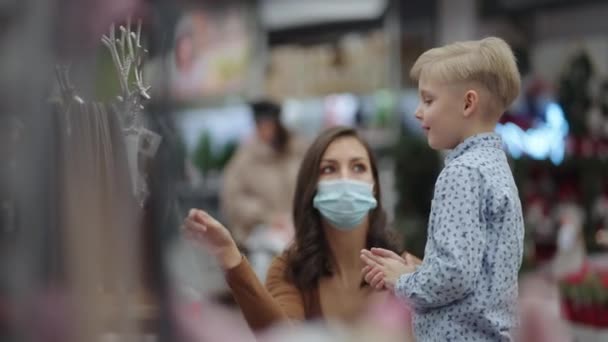 Mãe e filha vão às compras e escolhem árvores de Natal e brinquedos com decorações para o Natal em máscaras protetoras — Vídeo de Stock