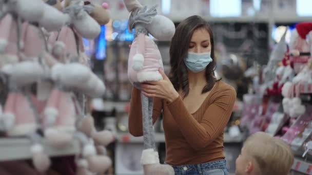 Koruyucu maskeli bir anne oyuncağı mağazadan alıp çocuğa gösterir. — Stok video