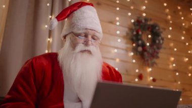 Noel Baba 'nın uzaktan çekilmiş bir fotoğrafı. Noel ağacının yanında dizüstü bilgisayarla koltukta oturuyor ve bilgisayarla çalışıyor.