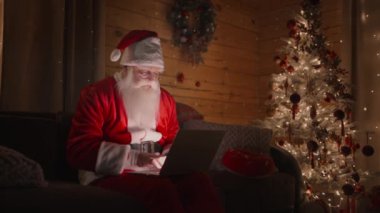 Evde Noel Baba 'nın portresi kanepeye oturmuş, dizüstü bilgisayarla Noel ağacının yanında çalışıyor.