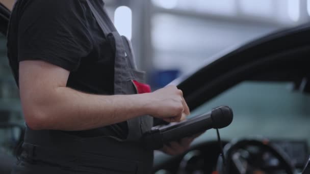 Un mecánico en un servicio de primer plano sostiene una tableta en sus manos y presiona la pantalla cerca del coche — Vídeos de Stock