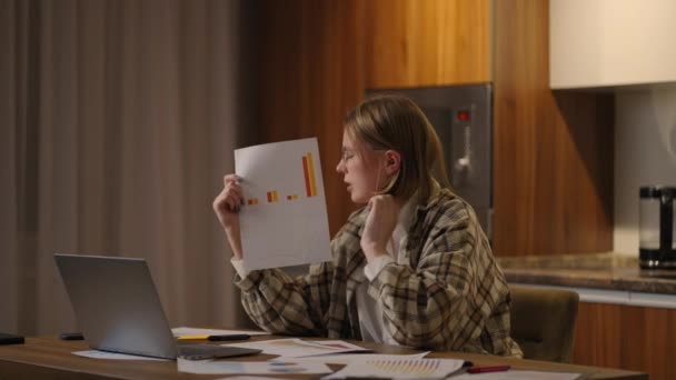 Bureau à domicile Une jeune femme portant des lunettes montre un graphique à une caméra d'ordinateur portable alors qu'elle était assise à la maison dans la cuisine et qu'elle faisait un reportage en ligne. Appel vidéo et conférence avec démonstration de documents — Video