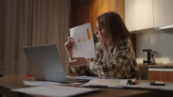 Evinde gözlüklü genç bir kadın, dizüstü bilgisayarında bir grafikle evde oturmuş online bir rapor gösteriyor. Belgeler ve programların gösterimi ve analizi ile video çağrısı ve konferansı — Stok video