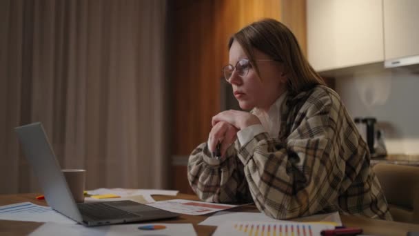 Uma mulher pensativa em óculos olha para a tela do laptop e se concentra e vem com uma estratégia de desenvolvimento. Horários e planos em papel estão na mesa — Vídeo de Stock