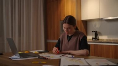 Yan bakış. Bir kadın bilgisayarlı bir masadaki ev ofisinde uzaktan çalışıyor ve bir grafikle ilgili verileri not alıyor, bir iş ekonomisti analisti. Not alır, not alır.