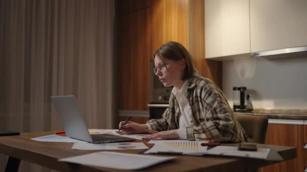 Zoom Una donna con gli occhiali lavora a distanza da casa seduta a un tavolo con un computer portatile e un pennarello segnala i dati sul grafico. A distanza studente a casa guarderà una lezione — Video Stock