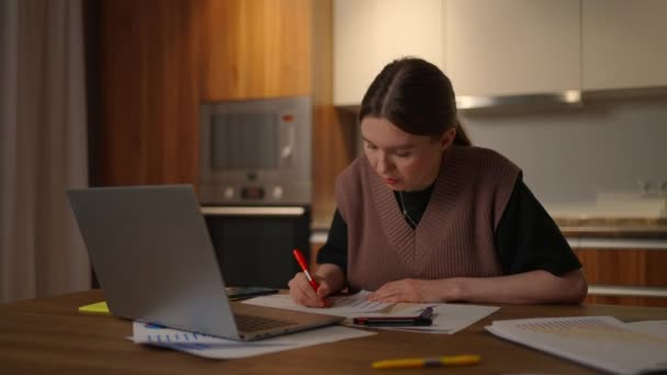 Retrato de uma mulher trabalhando remotamente em um escritório em casa em uma mesa com um laptop e notas de dados sobre um gráfico, analista economista de negócios — Vídeo de Stock