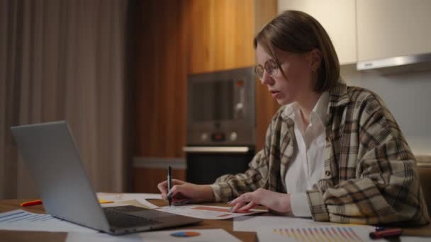 Retrato de uma mulher com óculos trabalhando remotamente em um escritório em casa em uma mesa com um laptop e notas de dados sobre um gráfico, economista de negócios — Vídeo de Stock