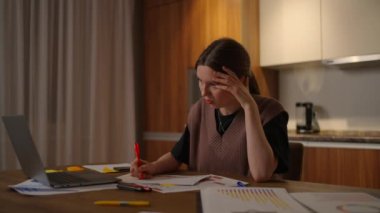 Yan bakış. Bir kadın bilgisayarlı bir masadaki ev ofisinde uzaktan çalışıyor ve bir grafikle ilgili verileri not alıyor, bir iş ekonomisti analisti. Not alır, not alır. Uzaklık öğrencisi ekonomist