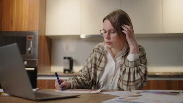 En kvinna arbetar på distans hemifrån sittandes vid ett bord med en bärbar dator och en filtspets penna markerar data på grafen. Distansstuderande hemma för att utföra ett uppdrag i ekonomi — Stockvideo