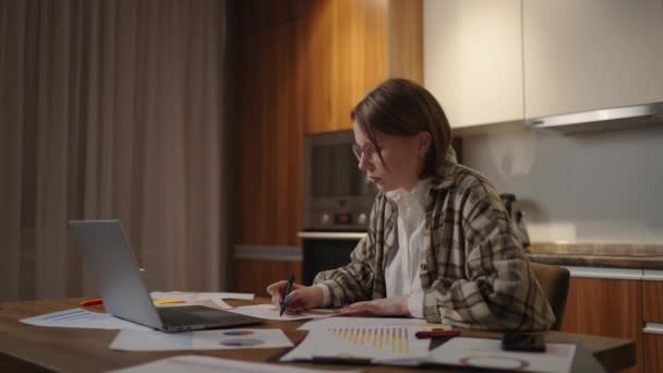 Eine Frau mit Brille arbeitet von zu Hause aus an einem Tisch sitzend mit einem Laptop und einem Filzstift, der die Daten auf der Grafik markiert. Fernstudentin schaut sich zu Hause Vorlesung an — Stockvideo