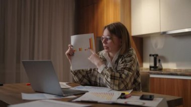 Evde gözlüklü genç bir kadın, evde dizüstü bilgisayarında bir grafik gösteriyor. Mutfakta oturmuş, online bir rapor hazırlıyor..