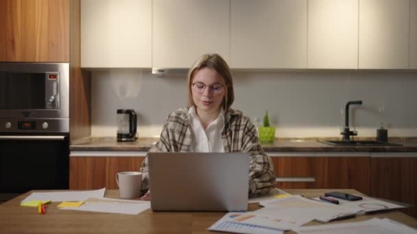 Cámara de zoom Una mujer con gafas trabaja remotamente desde casa sentada en una mesa con un portátil y un rotulador marca los datos en el gráfico. Estudiante de educación a distancia en casa para realizar una tarea — Vídeos de Stock