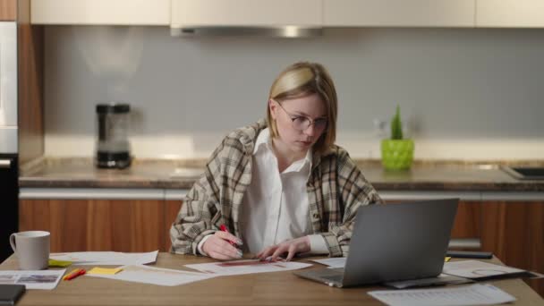 Une femme avec des lunettes travaille à distance de la maison assise à une table avec un ordinateur portable et un stylo feutre marque les données sur le graphique. Étudiant à distance à domicile pour effectuer une mission en économie — Video