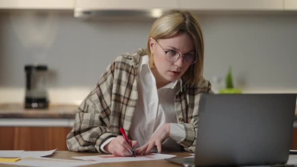 A woman with glasses works remotely from home sitting at a table with a laptop and a felt-tip pen marks the data on the graph. — Stock Video