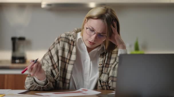 La mujer trabaja remotamente desde casa sentada en una mesa con un portátil y un rotulador marca los datos en el gráfico. Nerviosamente hablando en el enlace de vídeo — Vídeo de stock