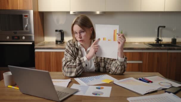 A woman with glasses at home shows a graph to a laptop camera while sitting at home in the kitchen. Home office remote work. Video call and conference with graphs and tables. — Stockvideo